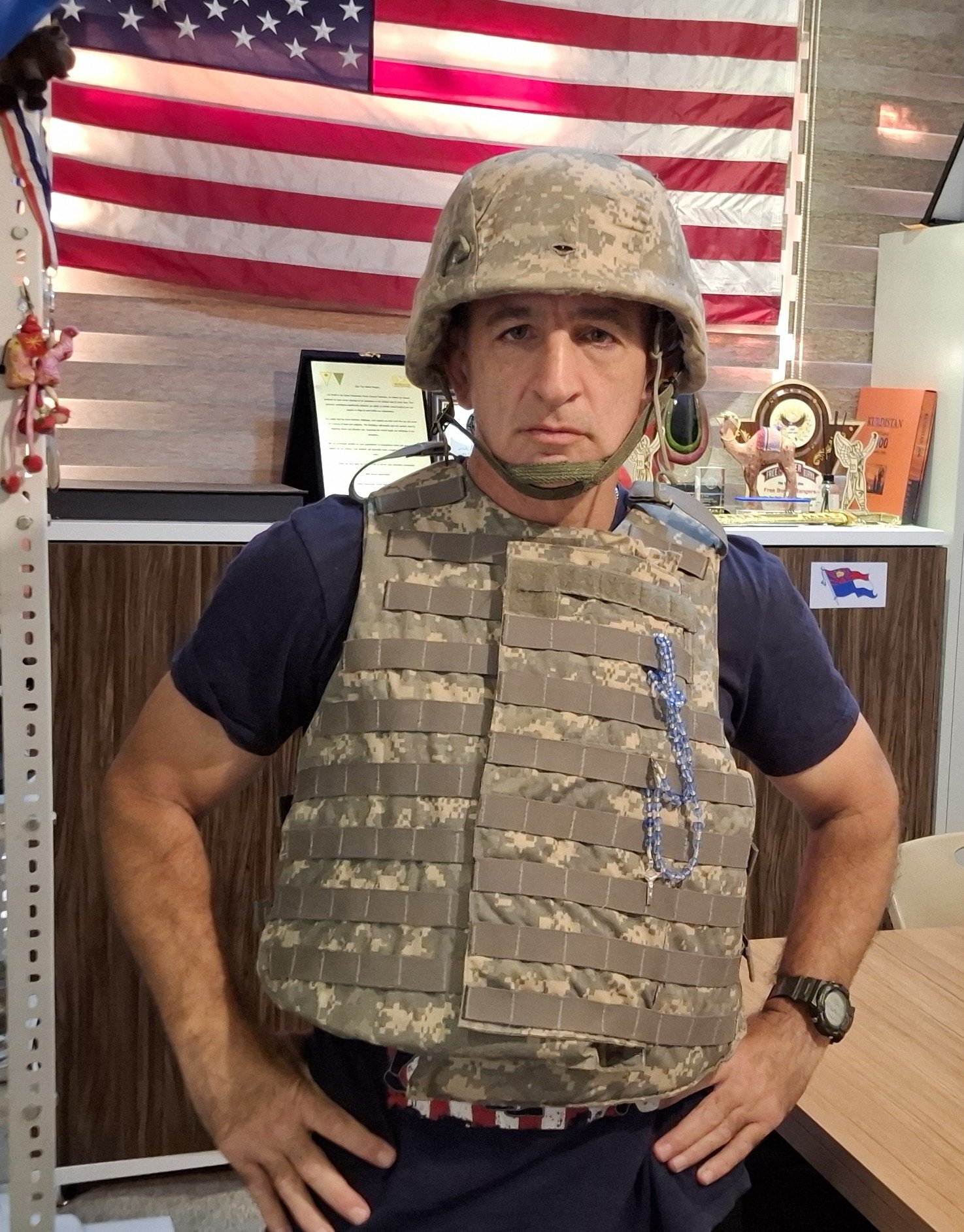 Man in military gear, including a camouflage vest and helmet, stands confidently in front of an American flag backdrop, showcasing a patriotic environment.