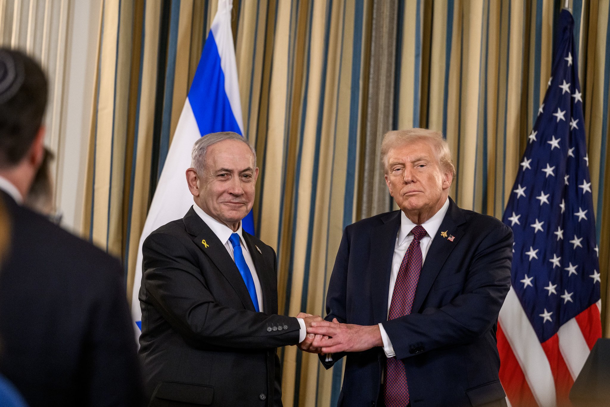 Israeli Prime Minister Benjamin Netanyahu and former U.S. President Donald Trump shake hands during a formal event, with flags of both nations in the background.